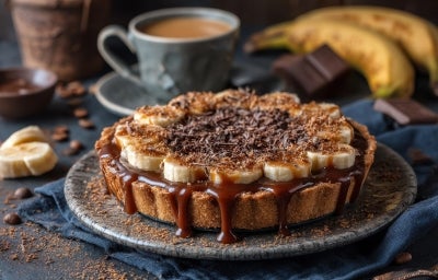 Em cima de uma mesa, prato com banoffee com massa de biscoitos crocante, bananas em rodelas, polvilhado com chocolate em pó.