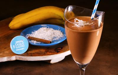 Fotografia em tons de azul em uma bancada de madeira, uma tábua de madeira com um copo de vidro alto e a bebida de chocolate com coco e banana dentro dele. Ao fundo, um prato azul com raspas de coco, uma canela em pau e uma banana. Fotografia em tons de azul em uma bancada de madeira, uma tábua de madeira com um copo de vidro alto e a bebida de chocolate com coco e banana dentro dele. Ao fundo, um prato azul com raspas de coco, uma canela em pau e uma banana.