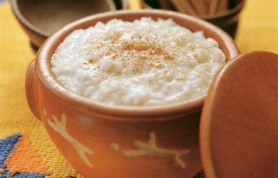 Fotografia em tons de amarelo em uma mesa de madeira com uma toalha amarela desenhada, um recipiente de barro com o arroz doce dentro dele. Ao fundo, um potinho com paus de canela. Fotografia em tons de amarelo em uma mesa de madeira com uma toalha amarela desenhada, um recipiente de barro com o arroz doce dentro dele. Ao fundo, um potinho com paus de canela.