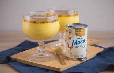 Foto da receita de Mousse de maracujá feita com o suco concentrado da fruta, servida em taças de vidro, com uma decoração de calda de maracujá com sementes, sobre uma tábua de madeira e ao lado de uma lata de Leite Moça