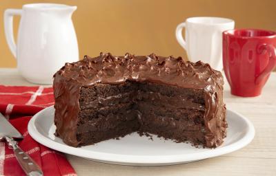 Fotografia em tons de vermelho em uma bancada de madeira com um pano vermelho listrado em branco, com um prato branco no centro com o bolo brownie com brigadeiro de cacau de cobertura. Ao fundo, uma xícara branca, uma xícara vermelha e um bule branco. Fotografia em tons de vermelho em uma bancada de madeira com um pano vermelho listrado em branco, com um prato branco no centro com o bolo brownie com brigadeiro de cacau de cobertura. Ao fundo, uma xícara branca, uma xícara vermelha e um bule branco.
