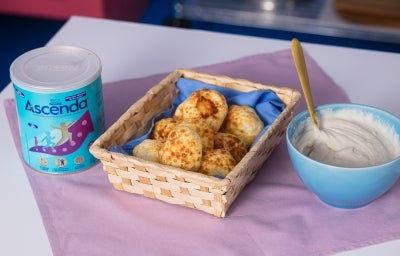 Fotografia em tons claros de uma cestinha de madeira retangular sobre um pano quadrado roxo. Dentro da cestinha tem um paninho menor azul com os pães de queijo, ao lado tem uma lata de Ascenda e um recipiente azul fundo pequeno com o creme de inhame.