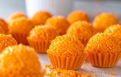 Fotografia mostra brigadeiros laranjas em forminhas laranja sobre uma mesa de madeira.