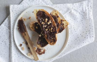 Fotografia vista de cima de algumas fatias de pão empilhadas com creme de avelã e chocolate em cada, em um prato branco raso. Ao lado, uma faca com um pouco do creme. O prato está em cima de um pano branco com pontos pretos, sobre uma mesa branca. Fotografia vista de cima de algumas fatias de pão empilhadas com creme de avelã e chocolate em cada, em um prato branco raso. Ao lado, uma faca com um pouco do creme. O prato está em cima de um pano branco com pontos pretos, sobre uma mesa branca.