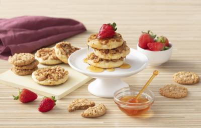Foto de uma bancada de madeira clara. Sobre ela há morangos soltos e em um pote, cookies em uma tábua, um pote de vidro com mel e um guardanapo vinho. Ao centro há um prato com três pancakes cobertas com mel e decoradas com morangos. Foto de uma bancada de madeira clara. Sobre ela há morangos soltos e em um pote, cookies em uma tábua, um pote de vidro com mel e um guardanapo vinho. Ao centro há um prato com três pancakes cobertas com mel e decoradas com morangos.