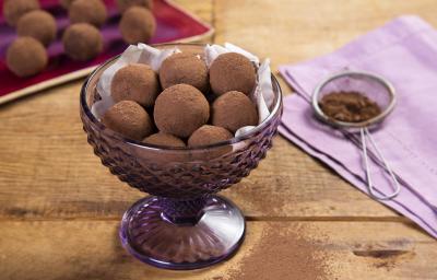 fotografia em tons de marrom e lilás de uma bancada marrom vista de cima, contém uma taça lilás com trufas redondas de chocolate e ao lado um pano lilás com uma peneira que contém cacau em pó.