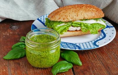 Fotografia de uma mesa de madeira e, sobre ela, um pote de vidro pequeno de vidro com pesto dentro. Ao lado tem u prato branco com detalhes azuis, apoiado no prato tem um sanduíche de frango com um pouco do pesto de coentro.