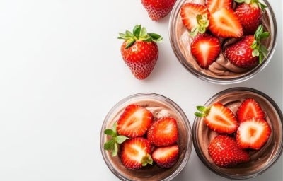 Fotografia em tons claros, vista de cima, de três recipientes de vidro pequenos com a sobremeza dentro, finalizados com ganache e morangos cortados pela metade. Os potinhos estão sobre uma mesa branca, ao lado dos potinhos tem morangos inteiros. Fotografia em tons claros, vista de cima, de três recipientes de vidro pequenos com a sobremeza dentro, finalizados com ganache e morangos cortados pela metade. Os potinhos estão sobre uma mesa branca, ao lado dos potinhos tem morangos inteiros.