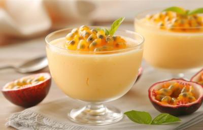 Fotografia de dois recipientes pequenos de vidro com mousse de maracujá e a polpa por cima. Os potinhos estão sobre uma mesa clara, com várias metades de maracujá.