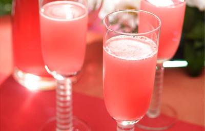 Fotografia em tons de rosa em uma bancada de madeira, um pano vermelho, taças de vidro de champagne com as bebidas feitas com Picolé de Limão com espumante.