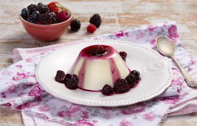 fotografia em tons de branco e roxo, contém um pano branco com rosas desenhadas, acima um prato redondo branco com um mini Flan com calda de frutas vermelhas e frutas vermelhas em volta.