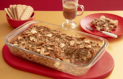 Foto da receita de Crumble de Pera. Observa-se um recipiente de vidro com o crumble sobre uma tábua vermelha. Atrás, um pratinho com uma porção e ao lado um copo de cappuccino.