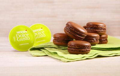 Fotografia em tons de verde em uma bancada de madeira clara, um paninho verde com vários macarons de chocolate e café em cima dele. Ao lado, cápsulas de Cappuccino Dolce Gusto. Fotografia em tons de verde em uma bancada de madeira clara, um paninho verde com vários macarons de chocolate e café em cima dele. Ao lado, cápsulas de Cappuccino Dolce Gusto.