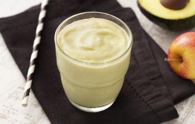 Fotografia em tons de branco e preto de uma bancada de madeira branca com um paninho preto, sobre ele um copo de vidro com o creme de abacate e um canudo. Ao fundo meio mamão e uma maçã. Fotografia em tons de branco e preto de uma bancada de madeira branca com um paninho preto, sobre ele um copo de vidro com o creme de abacate e um canudo. Ao fundo meio mamão e uma maçã.
