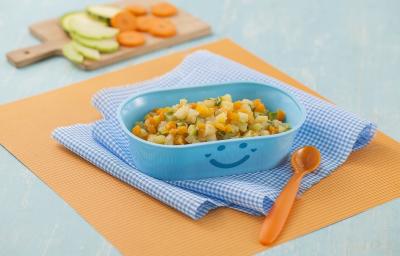 Fotografia em tons de laranja em uma bancada de madeira de cor azul. Ao centro, um pano laranja e um pano quadriculado azul, em cima, há um recipiente contendo os legumes e ao lado, uma colher laranja. Ao fundo, uma tábua contendo alguns legumes.