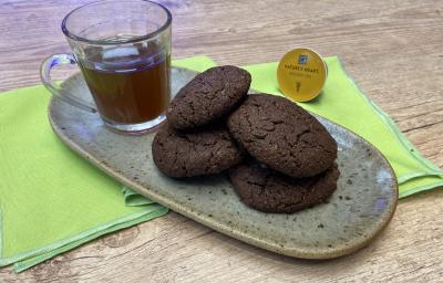 Fotografia em tons de verde em uma bancada de madeira, um recipiente oval marrom ao centro com 4 cookies de chocolate com amêndoas e ao lado uma xícara de vidro com o chá mate com acerola. Ao lado, uma cápsula de Dolce Gusto.