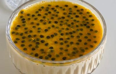 Foografia em tom claro de um recipiente de vidro decorativo sobre uma mesa branca. Dentro do recipiente tem uma receita de mousse de maracujá com calda de maracujá por cima.