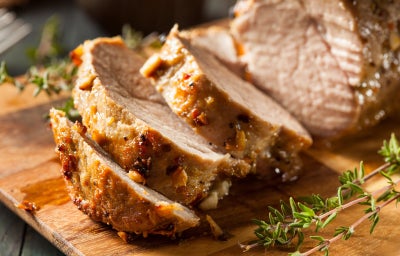Imagem de um lombo assado fatiado sobre uma tábua e ramos de tempero ao lado.