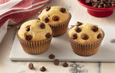 Fotografia em tons de branco e vermelho de uma bancada branca com uma tábua banca, sobre ela três muffins. Ao fundo um paninho vermelho e um recipiente redondo vermelho com gotas de chocolate.