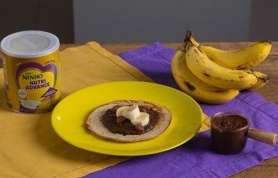 Fotografia de uma mesa de madeira com dois panos, roxo e amarelo, um por cima do outro. Apoiado neles tem um prato amarelo com uma panqueca de banana e creme de avelã. Ao redor tem uma lata de NINHO Nutri Advance, um cacho de bananas e o creme.