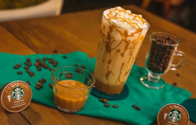 Fotografia em tons de verde de uma mesa de madeira com um paninho verde e ao centro um copo decorado com doce de leite, com uma bebida de leite e decorado com chantilly. Ao redor cápsula e grãos de café.