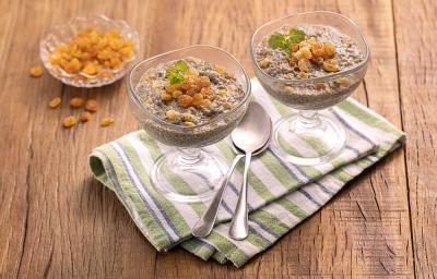 Foto da receita de Pudim de Chia com Uva Passa. Observa-se duas taças de vidro transparentes com o pudim e as uvas, sobre um guardanapo listrado em tons de verde claro. Também há duas colheres sobre o guardanapo e, atrás, há um pote com uvas passas.