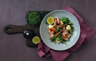 Fotografia vista de cima de um prato fundo cinza com uma salada de alface e agrião, presunto cru e vinagrete. O prato está apoiado sobre um pano rosa de tom escuro, que está em cima de uma tábua preta.