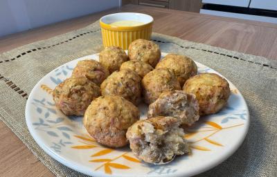 Fotografia em tons claros de um prato raso médio branco sobre um paninho em tom pastel sobre uma mesa de madeira. Dentro do prato tem várias unidades do bolinho de atum com batata, e dois deles na frente estão pela metade. Fotografia em tons claros de um prato raso médio branco sobre um paninho em tom pastel sobre uma mesa de madeira. Dentro do prato tem várias unidades do bolinho de atum com batata, e dois deles na frente estão pela metade.