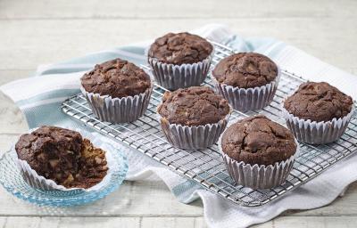 Fotografia em tons de azul em uma mesa de madeira clara com um suporte de ferro com os cupcakes integrais de cacau e banana em forminhas de papel. Ao lado, um pratinho de vidro com um cupcake cortado ao meio. Fotografia em tons de azul em uma mesa de madeira clara com um suporte de ferro com os cupcakes integrais de cacau e banana em forminhas de papel. Ao lado, um pratinho de vidro com um cupcake cortado ao meio.