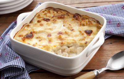Fotografia em tons de azul em uma bancada de madeira, um pano azul listrado, um recipiente quadrado branco com o frango gratinado dentro dele. Ao lado, uma colher. Fotografia em tons de azul em uma bancada de madeira, um pano azul listrado, um recipiente quadrado branco com o frango gratinado dentro dele. Ao lado, uma colher.