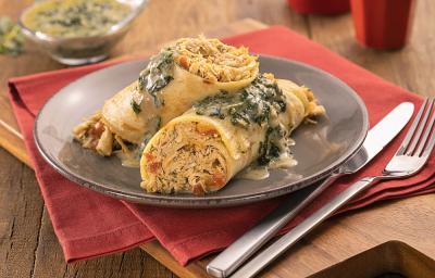 Foto em tons de amarelo da receita de panqueca de frango com molho de espinafre servida em duas porções em um prato cinza sobre um pano vermelho com talheres prateados ao lado. Tudo sobre uma tábua de madeira com uma recipiente ao fundo com o molho Foto em tons de amarelo da receita de panqueca de frango com molho de espinafre servida em duas porções em um prato cinza sobre um pano vermelho com talheres prateados ao lado. Tudo sobre uma tábua de madeira com uma recipiente ao fundo com o molho