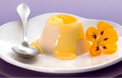 foto em tons lilas e branco. Acima de um prato roxo contém um prato na cor branco que comporta Flan. Ao lado uma colher e uma flor para decorar.o