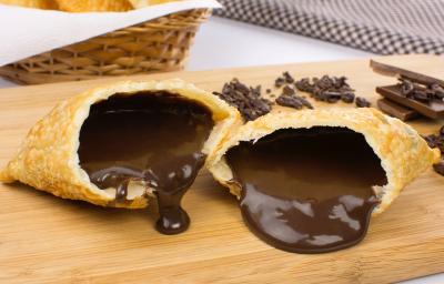 Fotografia em tons de amarelo em uma bancada de madeira de cor branca. Ao centro, há uma tábua de madeira contendo os pasteis e ao fundo há alguns chocolates espalhados e um pano quadriculado preto.