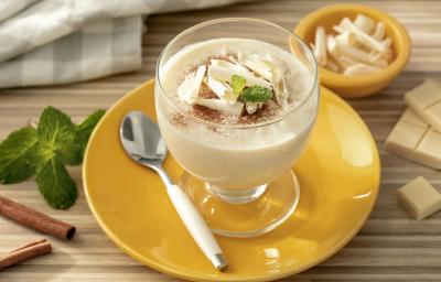 Fotografia em tons de branco em uma bancada de madeira de cor marrom. Ao centro, um pires amarelo contendo uma taça com o arroz doce. Ao lado, há uma colher e alguns pedaços de chocolate, canela e folha de hortelã. Ao fundo, um pano quadriculado cinza.