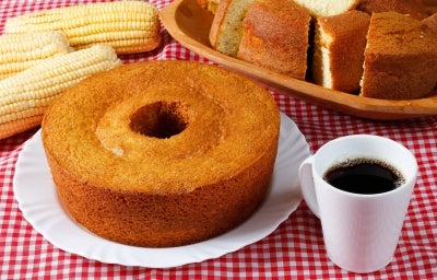 Fotografia de um bolo de milho com coco, exibido em um prato branco sobre uma toalha de mesa xadrez vermelha e branca. O bolo, com crosta dourada, é acompanhado por espigas de milho e uma caneca de café. Fotografia de um bolo de milho com coco, exibido em um prato branco sobre uma toalha de mesa xadrez vermelha e branca. O bolo, com crosta dourada, é acompanhado por espigas de milho e uma caneca de café.