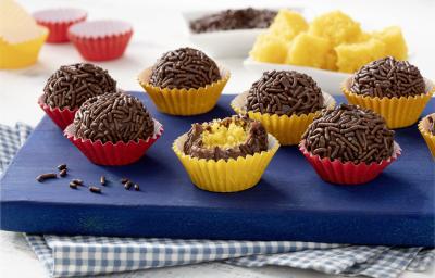 Fotografia em tons de azul, vermelho, amarelo e marrom de uma bancada com um pano quadriculado azul e branco, uma tábua azul, com 7 brigadeiros em cima. Ao centro um brigadeiro mordido, mostrando o recheio de bolo de cenoura.