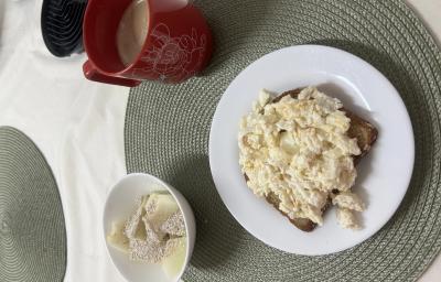 Fotografia de um prato raso médio branco com uma fatia de pão e ovos mexidos cremosos com farelo de aveia. Ao lado tem um recipiente pequeno de vidro e branco com melão e aveia e uma xícara vermelha grande. Fotografia de um prato raso médio branco com uma fatia de pão e ovos mexidos cremosos com farelo de aveia. Ao lado tem um recipiente pequeno de vidro e branco com melão e aveia e uma xícara vermelha grande.