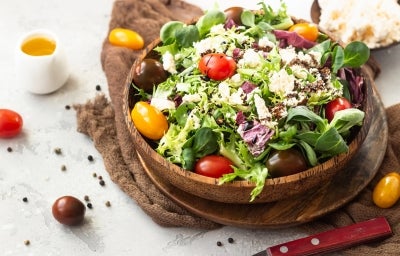 Fotografia de uma salada vibrante em uma tigela, com uma mistura de folhas verdes, tomates-cereja em várias cores e queijo branco esfarelado, regada com molho balsâmico. A composição tem um recipiente de azeite e grãos de pimenta.