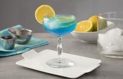 Foto em tons de azul da receita do drink Lagoa Azul servida em uma taça de vidro alta sobre uma base de porcelana branca. Ao fundo há um copo de vidro com cubos de gelo, além de laranjas cortadas e um pano azul para decoração