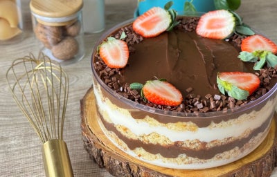 Fotografia mostra um bolo em camadas de creme branco, massa e chocolate. O bolo está coberto com uma camada de chocolate.