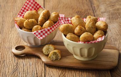 Fotografia em tons de marrom, com dois bowls de porcelana forrados com guardanapos quadriculados em vermelho e branco, sendo um sobre tábua com um bolinho aberto, ambos com bolinhos de macaxeira fritos inteiros, tudo sobre bancada de madeira rústica. Fotografia em tons de marrom, com dois bowls de porcelana forrados com guardanapos quadriculados em vermelho e branco, sendo um sobre tábua com um bolinho aberto, ambos com bolinhos de macaxeira fritos inteiros, tudo sobre bancada de madeira rústica.