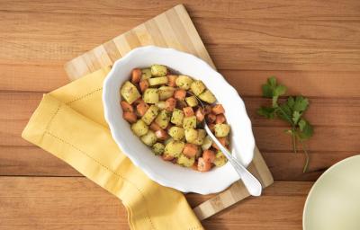 Fotografia em tons de branco, amarelo e marrom, ao centro porcelana branca com legumes cozidos variados polvilhados com coentro picado e uma colher, guardanapo listrado bege e outro amarelo, lado direito prato bege e ramo de coentro, em bancada marrom.