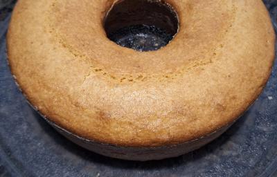 Fotografia de um bolo de fubá, feito na forma de furo central, apoiado em um vidro redondo, sobre uma bancada de cor escura. Fotografia de um bolo de fubá, feito na forma de furo central, apoiado em um vidro redondo, sobre uma bancada de cor escura.