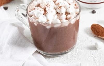 Fotografia em tons de marrom em uma mesa de madeira branca, um pano branco e uma xícara de vidro com o chocolate quente dentro e minis marshmallow para decorar a xícara. Ao lado, uma colherzinha de madeira. Fotografia em tons de marrom em uma mesa de madeira branca, um pano branco e uma xícara de vidro com o chocolate quente dentro e minis marshmallow para decorar a xícara. Ao lado, uma colherzinha de madeira.
