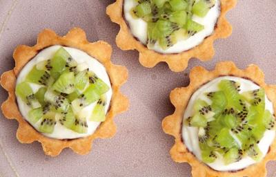 fotografia em tons de lilás, branco e verde tirada de um prato lilás redondo visto de cima, contém 3 mini-torta com pedaços de kiwi e creme. fotografia em tons de lilás, branco e verde tirada de um prato lilás redondo visto de cima, contém 3 mini-torta com pedaços de kiwi e creme.