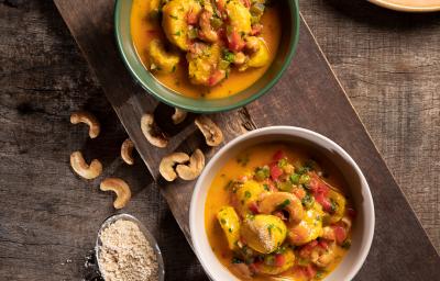 Foto de cima de dois pratos redondos e fundos com a receita de moqueca de banana da terra, ambos sobre uma tábua de madeira, com algumas castanhas-de-caju na mesa, uma colher de metal com arroz e um prato com arroz branco Foto de cima de dois pratos redondos e fundos com a receita de moqueca de banana da terra, ambos sobre uma tábua de madeira, com algumas castanhas-de-caju na mesa, uma colher de metal com arroz e um prato com arroz branco