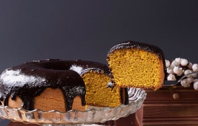 Apresenta o bolo de cenoura inteiro com uma fatia sendo retirada com uma espátula. A cobertura de chocolate está bem brilhante e espessa, escorrendo sobre o bolo. O fundo é escuro e elegante, destacando bem a cor dourada do bolo.