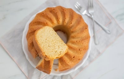 Fotografia de um bolo bundt recém-assado em um prato branco, com crosta dourada e interior fofinho, pronto para ser servido, acompanhado de dois garfos prateados em um ambiente acolhedor.