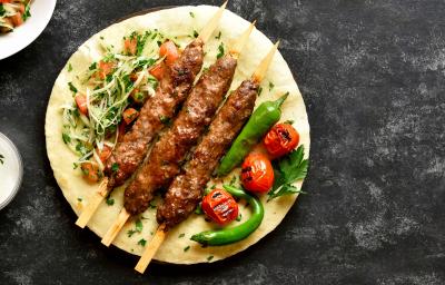 Fotografia de um pão redondo com 3 espetos de carne ao centro. Junto dos espetos de carne existe uma salada colorida acompanhada de algumas pimentas inteiras. Ao lado desse pão existe um pote pequeno com um molho branco. Fotografia de um pão redondo com 3 espetos de carne ao centro. Junto dos espetos de carne existe uma salada colorida acompanhada de algumas pimentas inteiras. Ao lado desse pão existe um pote pequeno com um molho branco.