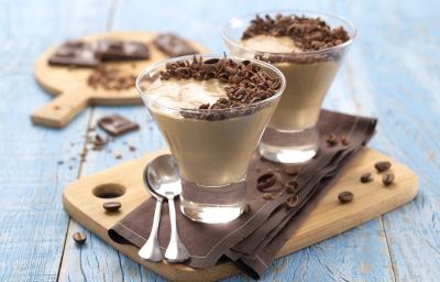 Foto da receita de mousse de cappuccino. Observa-se uma superfície de madeira azul-clara, em cima dela uma pequena tábua de madeira com um guardanapo marrom e duas taças de vidro com a mousse apoiadas. Como decoração, raspas de chocolate e grãos de café Foto da receita de mousse de cappuccino. Observa-se uma superfície de madeira azul-clara, em cima dela uma pequena tábua de madeira com um guardanapo marrom e duas taças de vidro com a mousse apoiadas. Como decoração, raspas de chocolate e grãos de café